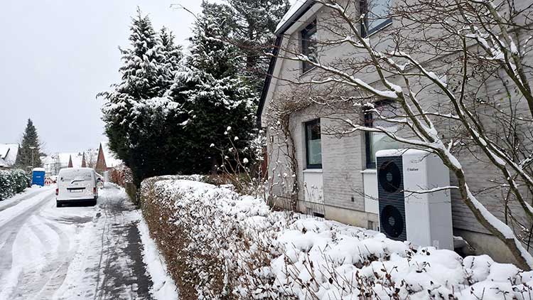 Straße in einem Wohngebiet und Haus, vor dem eine Luftwärmepumpe steht 