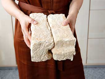 Eine Person hält zwei rechteckige Blöcke aus festem, cremefarbenem Material, die nach dem Nut-und-Feder-Prinzip zusammenpassen. Die Oberfläche der Blöcke ist unregelmäßig und weist eine faserige, strukturierte Textur auf. 
