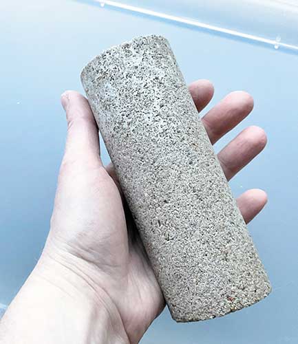Das Bild zeigt eine Hand, die einen Zylinder aus Biobeton von ca. 25 cm Länge hält.