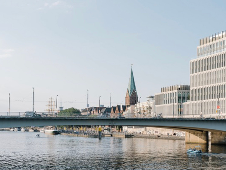 Städtische Landschaft mit Kirchturm mittig, modernen Gebäuden rechts, Brücke über ruhiges Wasser unter klarem Himmel. Fluss reflektiert Licht, verbindet historische und moderne Architektur. 