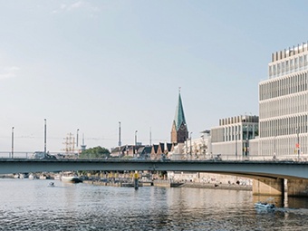 Städtische Landschaft mit Kirchturm miitig, modernen Gebäuden rechts, Brücke über ruhiges Wasser unter klarem Himmel. Fluss reflektiert Licht, verbindet historische und moderne Architektur. 
