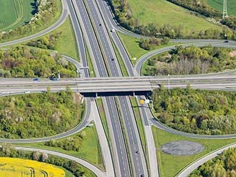 Luftaufnahme einer Autobahnbrücke mit den Ausfahrten von und den Auffahrten auf die Autobahn in einer stark begrünten Umgebung. 