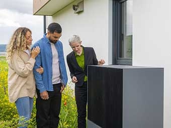 Eine Energieberaterin und ein junges Paar stehen vor einer Wärmepumpe. 