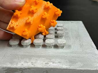 Das Bild zeigt ein Modell des Klettverschlusses zur Verbindung von Gebäudeteilen. Ein Teil ist eine graue Betonplatte mit pilzförmigen Noppen, der Gegenpart ist aus orangefarbigem Kunststoff und hat "Verbindungshaken" in Form des "Plus-Symbols" (ein Kreuz aus gleich langen Linien, die sich mittig schneiden). 