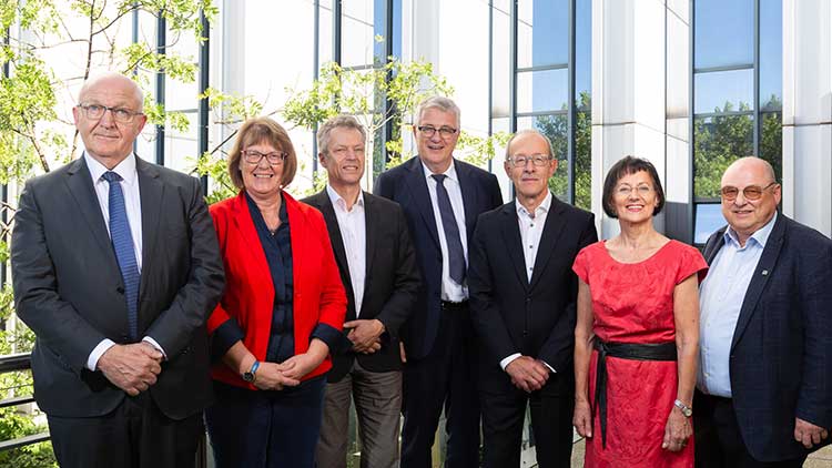 Eine Gruppe von sieben Personen – fünf Männer und zwei Frauen – in formeller Kleidung steht in zwei Reihen vor einem modernen Bürogebäude mit großen Glasfenstern. Im Hintergrund sind grüne Bäume zu sehen.  