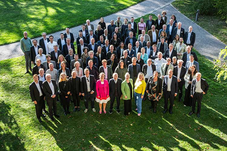 Gruppenbild mit etwa 90 Personen, überwiegend in businessorientierter Kleidung, aufgenommen aus erhöhter Perspektive im Freien. Die Menschen stehen auf einer Wiese, umgeben von Gehwegen und grüner Vegetation. Im Vordergrund stehen einige Personen in farbenfrohen Outfits (z. B. pink, gelb, grün). 