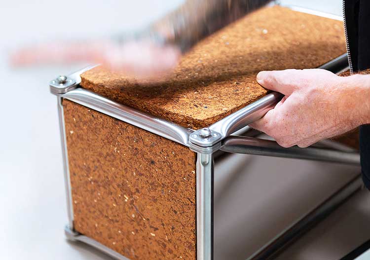 Nahaufnahme eines Regalmoduls aus Metall und Spanholz. Zwei Hände halten den Rahmen aus gebürstetem Metall, während die Spanplatte in den oberen Rahmen gepresst wird. Die Verbindungselemente an den Ecken sind durch Schrauben gesichert. 