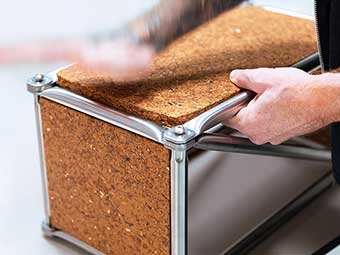 Nahaufnahme eines Regalmoduls aus Metall und Spanholz. Zwei Hände halten den Rahmen aus gebürstetem Metall, während die Spanplatte in den oberen Rahmen gepresst wird. Die Verbindungselemente an den Ecken sind durch Schrauben gesichert. 