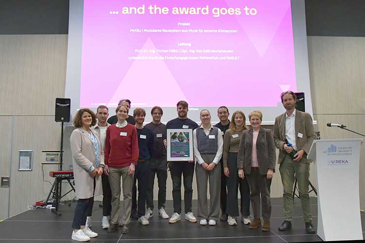 Gruppe von zwölf Personen steht auf einer Bühne vor einer projizierten Präsentation mit der Überschrift 