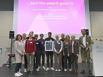 Gruppe von zwölf Personen steht auf einer Bühne vor einer projizierten Präsentation mit der Überschrift "... and the award goes to". Eine Person in der Mitte hält eine Urkunde. Im Hintergrund sind ein Rednerpult mit Logo der Universität und Lautsprecher sichtbar. 