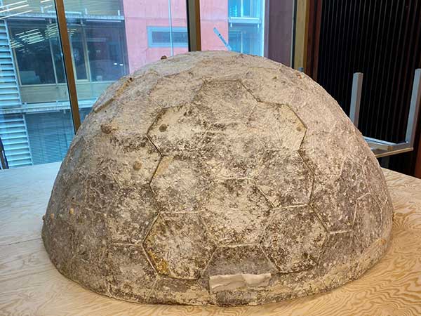 Halbkugelförmige Struktur aus einem erkennbar porösen Material mit hexagonalem Muster, das sich über die gesamte Oberfläche erstreckt. Die Konstruktion liegt auf einer Holzfläche, im Hintergrund sind Fenster mit Blick auf Gebäudefassaden aus Glas und farbigem Material zu sehen.