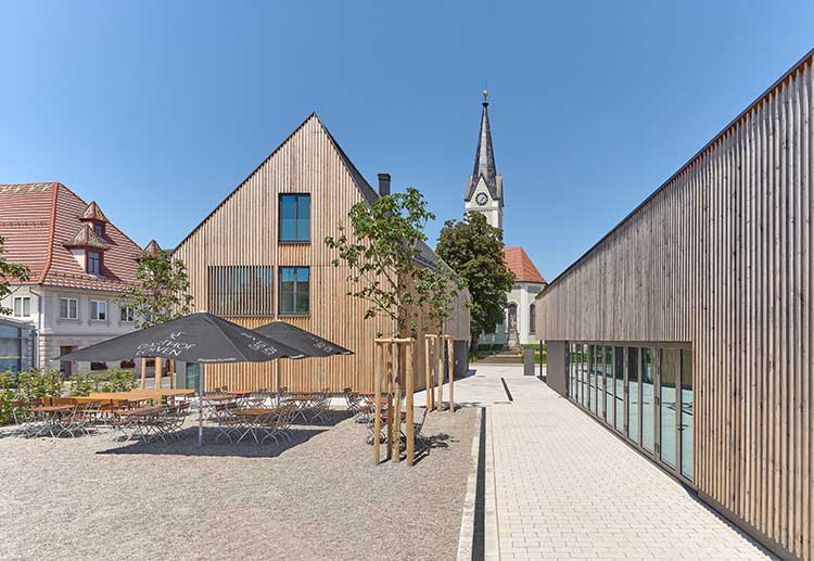 Moderner Gebäudekomplex mit Holzfassaden, bestehend aus zwei schmalen Baukörpern. Links ein Giebelhaus mit großflächigen Fenstern, rechts ein flacheres Gebäude mit Glasfront. 