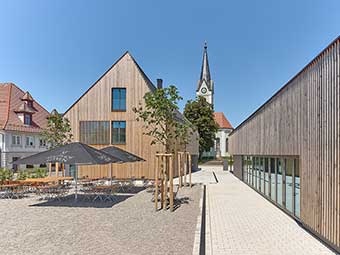 Moderner Gebäudekomplex mit Holzfassaden, bestehend aus zwei schmalen Baukörpern. Links ein Giebelhaus mit großflächigen Fenstern, rechts ein flacheres Gebäude mit Glasfront. 