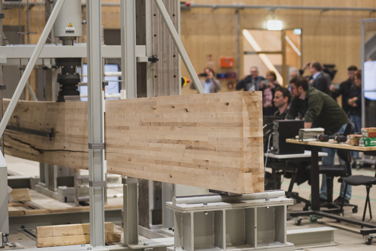 Foto: Holzbauteil in Prüfmaschine. Massiver Brettschichtholzbalken wird horizontal von einer vertikalen Druckpresse auf Belastung geprüft.