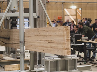 Foto: Holzbauteil in Prüfmaschine. Massiver Brettschichtholzbalken wird horizontal von einer vertikalen Druckpresse auf Belastung geprüft.  