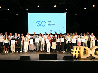 Gruppenfoto von mehreren Personen auf einer Bühne vor einer Leinwand mit dem Logo und Schriftzug „DGNB Sustainability Challenge 2025“. Die Personen stehen nebeneinander und halten Urkunden in den Händen. Rechts im Vordergrund steht eine leuchtende gelbe 