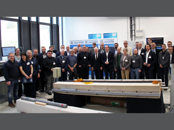 Eine Gruppe von etwa 20 Personen steht in einem hellen Raum mit großen Fenstern. Im Vordergrund ist ein Betonbauteil mit Sensorik-Elementen und Messgeräten auf einem Gestell zu sehen. 