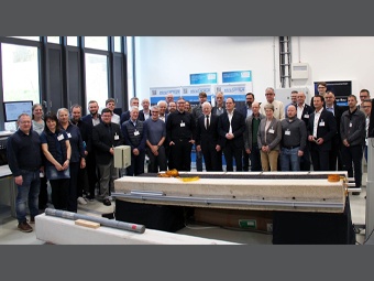 Eine Gruppe von etwa 20 Personen steht in einem hellen Raum mit großen Fenstern. Im Vordergrund ist ein Betonbauteil mit Sensorik-Elementen und Messgeräten auf einem Gestell zu sehen. 