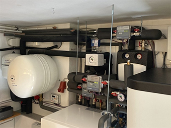 Heizungsanlage in einem Technikraum. Zu sehen sind ein großes, weißes Ausdehnungsgefäß und kleinere Komponenten wie Druckanzeigen, Absperrventile sowie ein Wärmemengenzähler. Schwarze Rohrleitungen verlaufen oben und seitlich, teilweise isoliert. Rechts ist ein Teil eines Pufferspeichers erkennbar. 