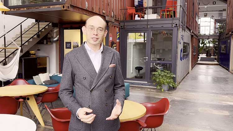 Eine Person in Business-Kleidung steht in einer modernen Büroumgebung, bestehend aus einer lichtdurchfluteten Halle mit Containern, die zu Arbeitsräumen umfunktioniert wurden.  
