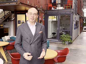 Eine Person in Business-Kleidung steht in einer modernen Büroumgebung, bestehend aus einer lichtdurchfluteten Halle mit Containern, die zu Arbeitsräumen umfunktioniert wurden.  