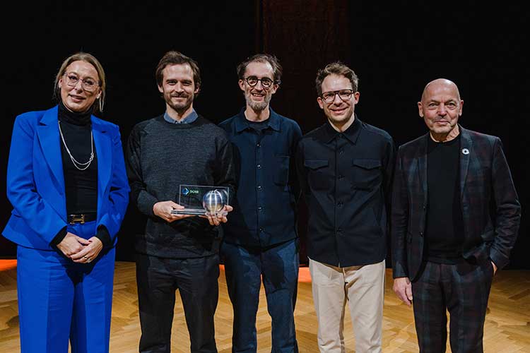 Fünf Personen in festlicher Kleidung vor einem schwarzen Hintergrund, eine hält eine Preisstatuette in der Hand. 