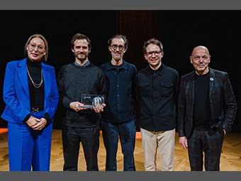 Fünf Personen in festlicher Kleidung vor einem schwarzen Hintergrund, eine hält eine Preisstatuette in der Hand. 