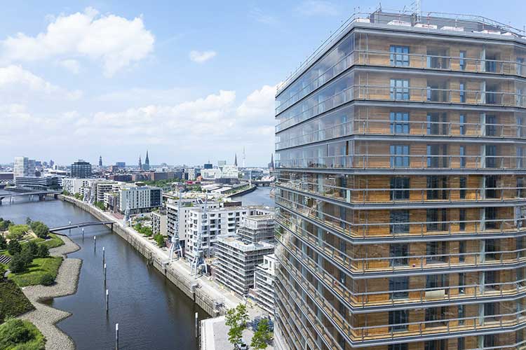 Modernes Hochhaus mit Holz-Glas-Fassade im Vordergrund rechts, umgeben von weiteren Wohn- und Bürogebäuden. Links ein Fluss mit Promenade und Brücken. Der Hintergrund zeigt eine urbane Skyline. 