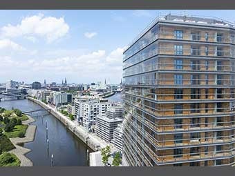 Modernes Hochhaus mit Holz-Glas-Fassade im Vordergrund rechts, umgeben von weiteren Wohn- und Bürogebäuden. Links ein Fluss mit Promenade und Brücken. Der Hintergrund zeigt eine urbane Skyline. 